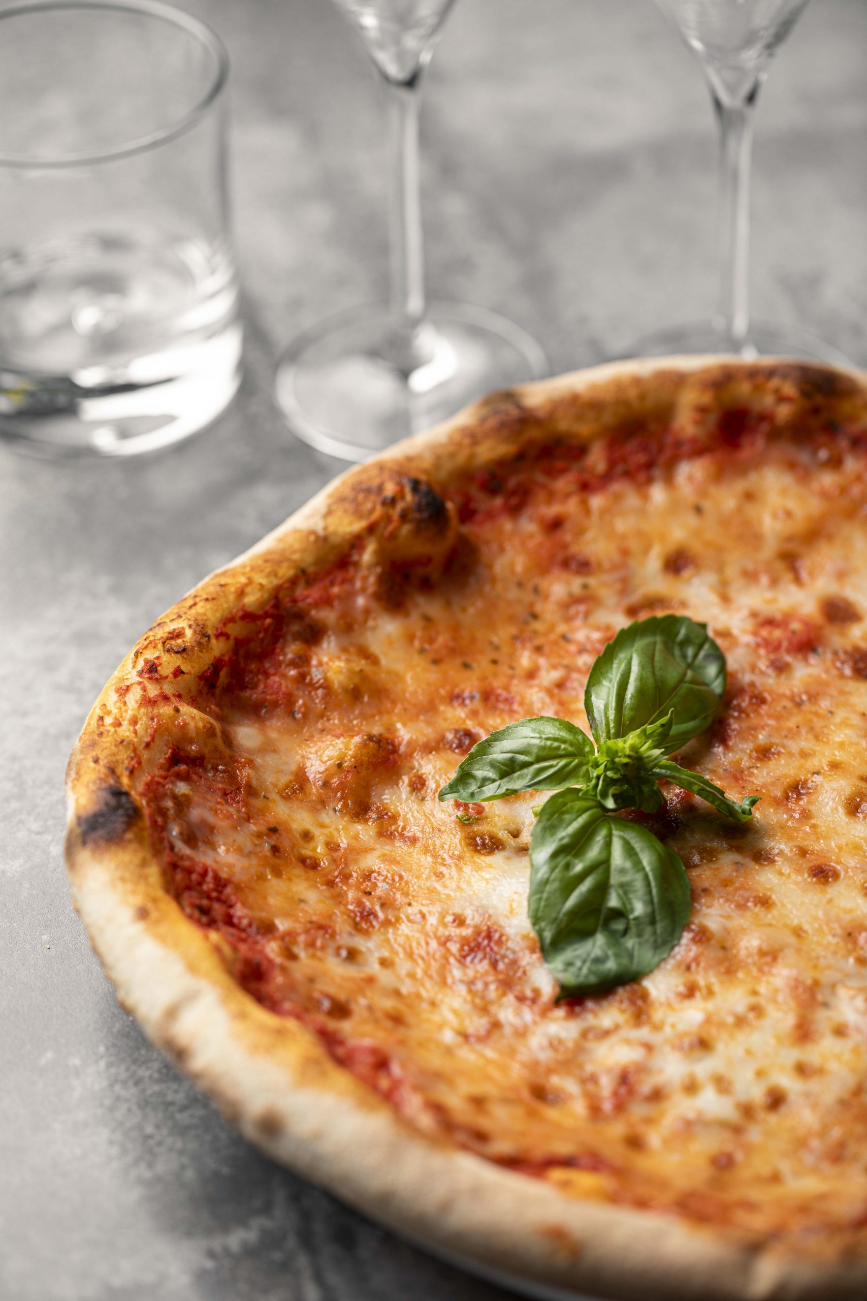 Pizza margherita maison proposée par le restaurant italien Mamma Mia à Echirolles Mamma Mia. La pizza est à la base de tomate avec une feuille de basilic. L'assiette est posée sur une table grise avec des verres en arrière plan.