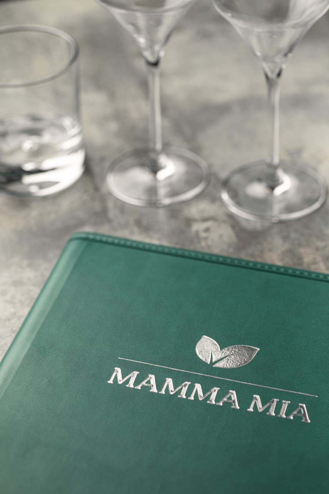Menu de restaurant Mamma Mia de couleur verte posée sur une table grise avec des verres en second plan.